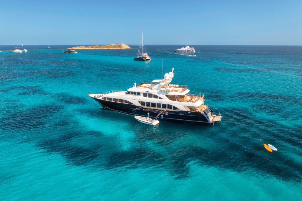 Aerial view of luxury yacht on blue sea at sunny day in summer. Sardinia, Italy. Aerial view of speed boats, yachts, sea lagoon, shore, transparent water, sky. Top view from drone. Tropical seascape Crewed Yacht Charter Vacations